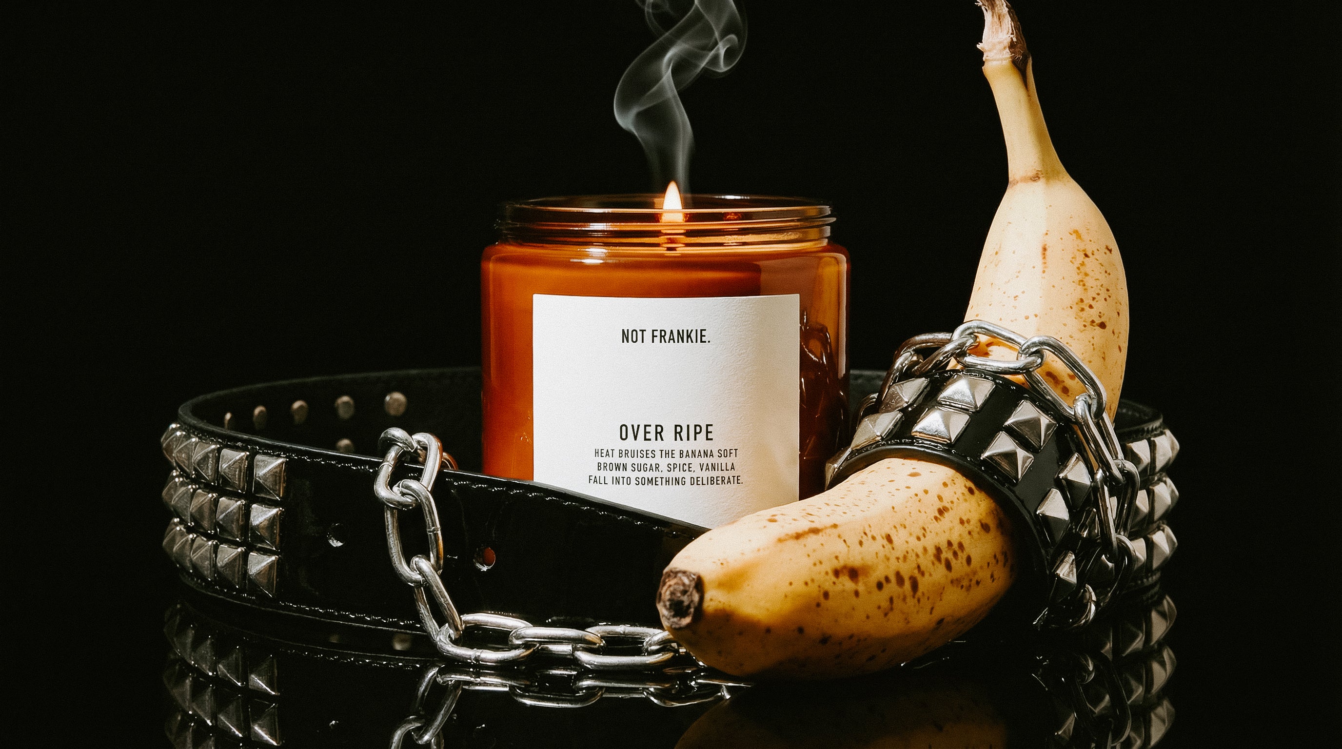 Not Frankie Over Ripe candle on a black lacquer surface, surrounded by a studded black leather belt and chrome chains, with a ripe speckled banana wrapped in spikes beside the candle. High-contrast, moody lighting with rising smoke from the flame
