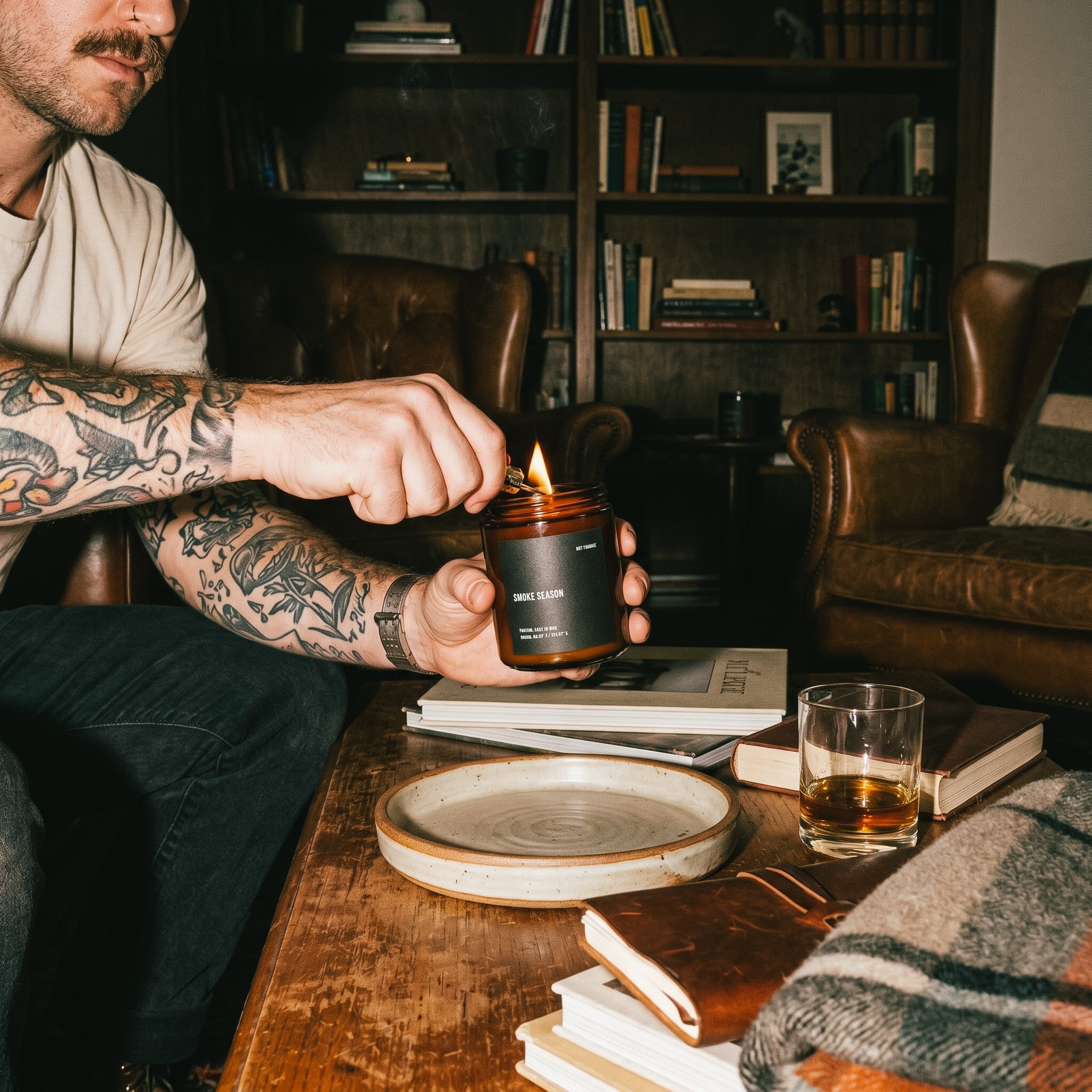 Tattooed person lighting a NOT FRANKIE Smoke Season candle in a moody lounge filled with books, leather chairs and warm ambient light, capturing the candle's smoky, woody winter atmosphere.