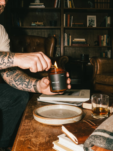 Tattooed person lighting a NOT FRANKIE Smoke Season candle in a moody lounge filled with books, leather chairs and warm ambient light, capturing the candle's smoky, woody winter atmosphere.